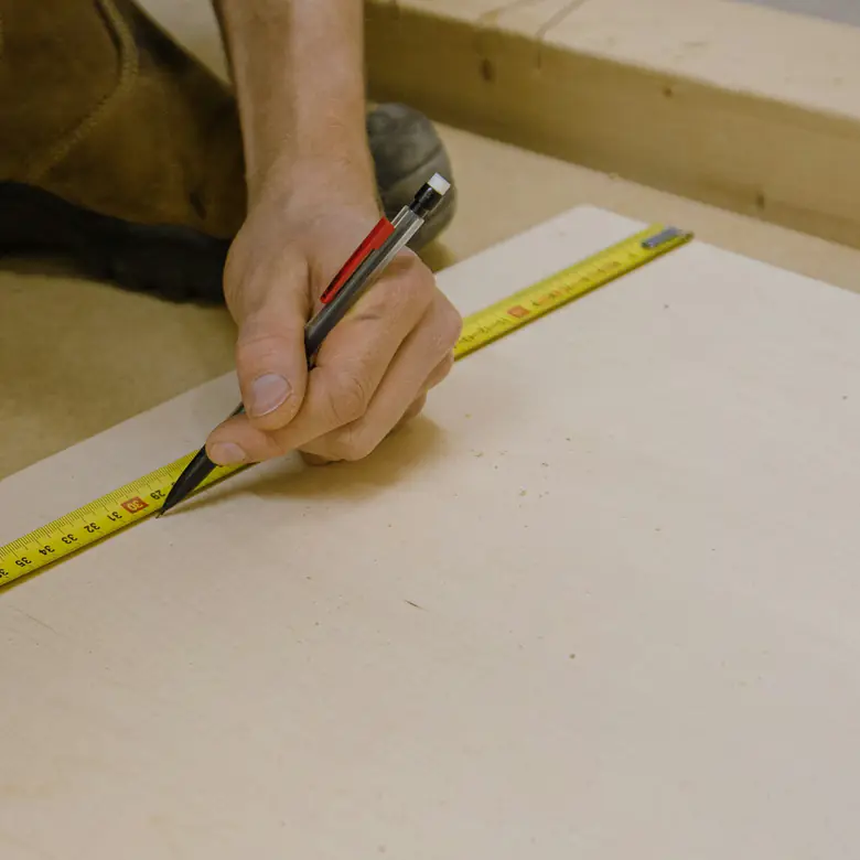 Photo d’illustration : une main reporte une mesure au crayon sur un panneau de bois, le long d’un mètre ruban.