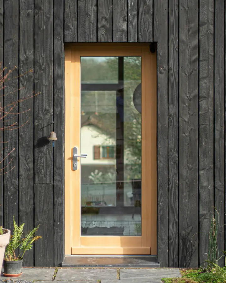 Photo d’illustration : une porte d’entrée vitrée en bois clair, dans une façade en bardage de bois noirci.